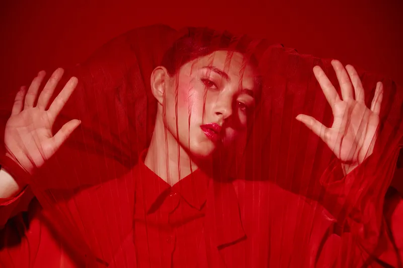 Woman in vibrant red outfit with sheer fabric covering face and hands, creating intricate shadows on smooth skin. Bold fashion and beauty concept with artistic lighting.