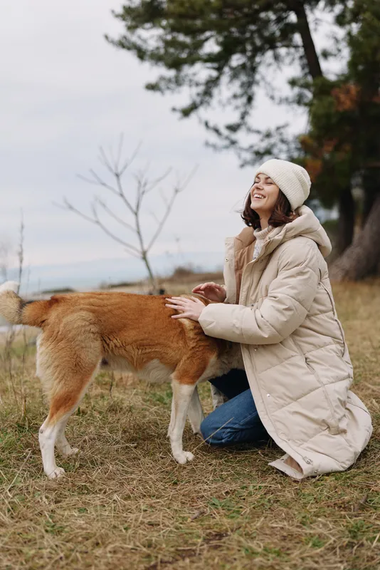 Woman kneeling in a beige coat and beanie outdoors, patting a friendly dog in an autumn field, calm moment of companionship and nature, soft light and gentle breeze, park scenery
