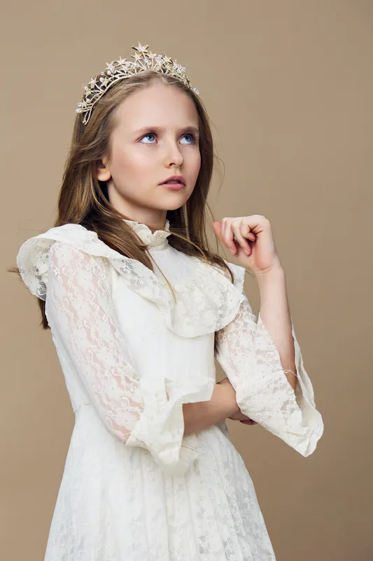 A young girl in a white lace dress with a crown, posing thoughtfully against a soft beige background, showcasing innocence and elegance in a seasonal concept