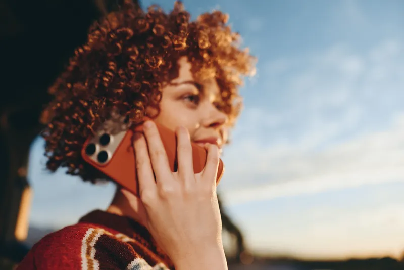 Woman lifestyle portrait outdoors at sunset, curly hair and face closeup, holding smartphone to her ear, casual knit sweater, communication by calling, relaxed hand gesture, soft evening sky