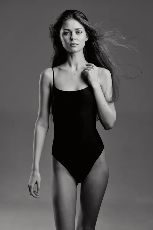 Black and white portrait of a woman in a black bodysuit with a serious expression on a gray background.