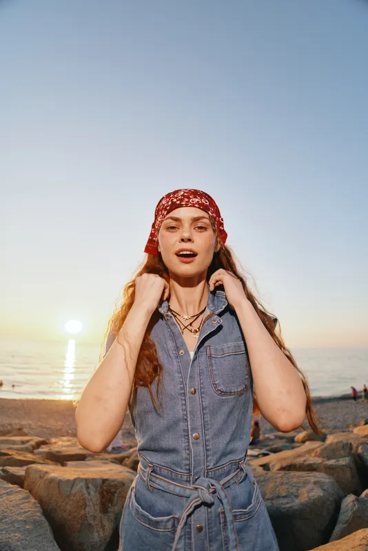 lifestyle woman in modern boho-western denim street style posing outdoors at sunset with warm film color and relaxed expression near rocky beach shore