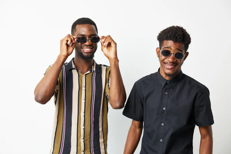 Two smiling African American men wearing stylish sunglasses pose confidently against a white background, showcasing modern fashion and friendship in a lively atmosphere.