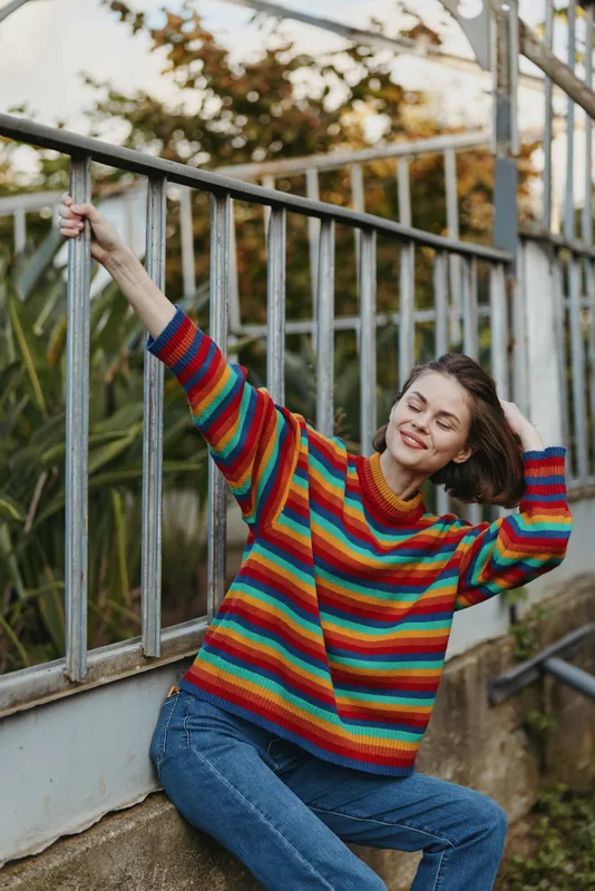 Woman in her mid 20s with short brown hair, wearing a colorful striped sweater and jeans, leaning on metal railing and smiling with closed eyes in a relaxed urban garden lifestyle portrait.