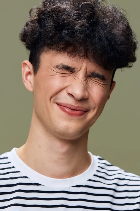 young man with curly hair squinting eyes and slight smile wearing striped shirt against green background in casual style, natural expression, close-up portrait