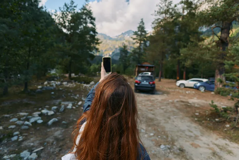 A person with long hair is seen taking a photo with a smartphone along a scenic mountain trail. They smile softly, appearing engaged and curious, creating an adventurous outdoor atmosphere.