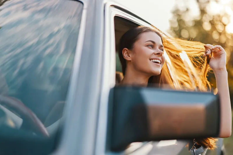 Joyful young woman enjoying a road trip with flowing hair, bright sunshine, and a scenic background, evoking freedom and adventure.