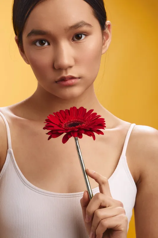 Asian woman holds a vibrant red flower close to her face, gazing softly at the camera, with a warm yellow background creating a sunny, inviting portrait.