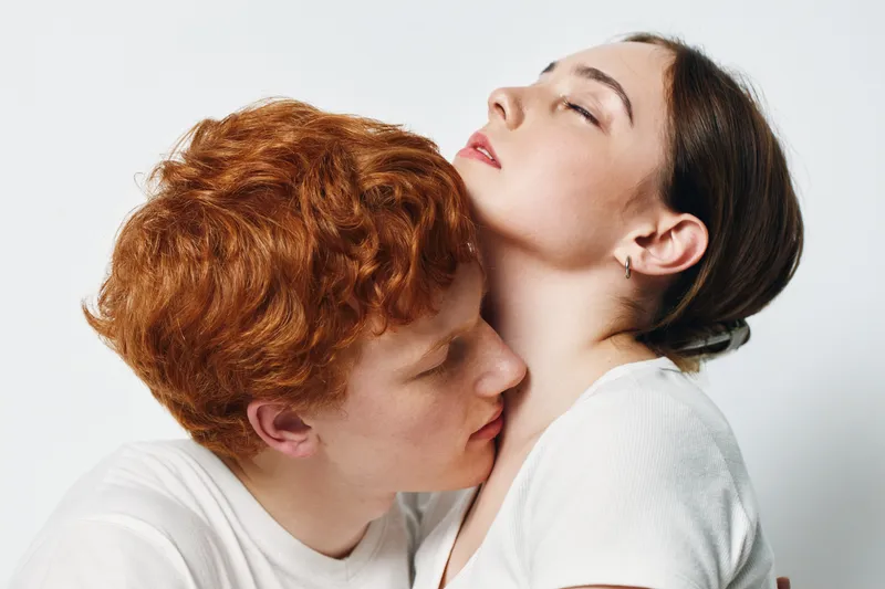 Couple, intimacy, affection and love with redhead man kissing woman neck in white clothes on light background. Young lovers with closed eyes sharing romantic moment and close connection together.