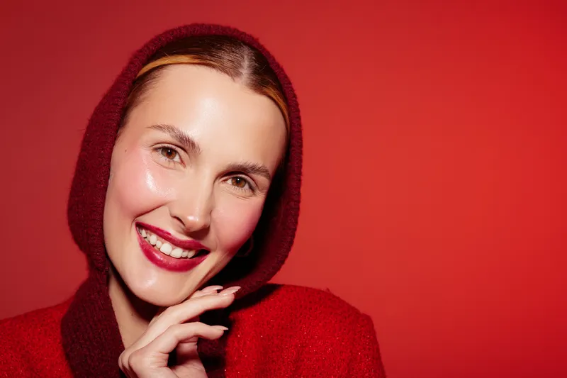 Smiling woman with shiny skin and red lipstick wears red hooded sweater against red background. Close-up portrait with bright makeup and joyful expression. Concept of beauty and fashion.