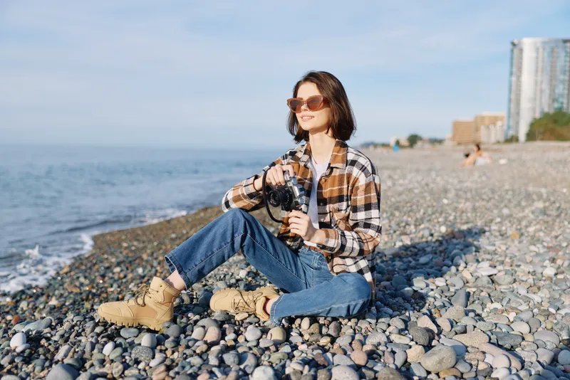 Beach scene with a woman in casual plaid shirt and jeans, sitting on pebbles along the shore, holding a camera and wearing sunglasses, enjoying the sunny coastal atmosphere.