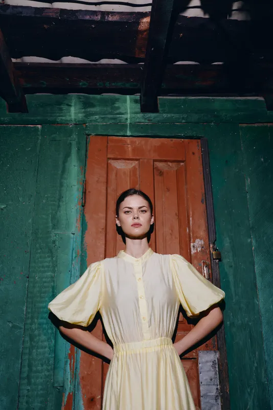 Woman portrait in vintage dress standing in doorway against rustic teal wall, confident pose hands on hips, authentic inclusive model, moody light, cinematic