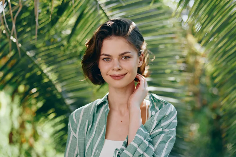 Portrait of a smiling young woman standing outdoors among tropical plants, wearing a green plaid shirt and natural makeup.