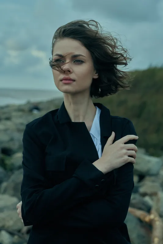 Windy coastline scene with a woman in a dark jacket gazing into the distance, sea spray in the air, moody sky, and rugged rocks creating a contemplative outdoor mood.