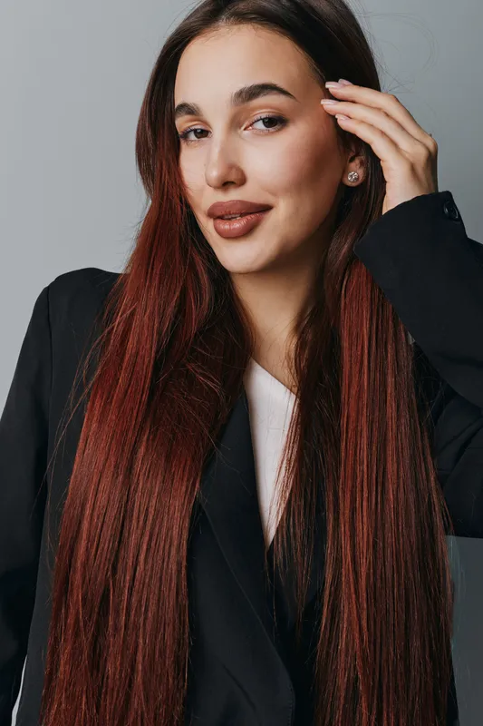 Woman with long straight red hair and natural makeup wearing black blazer and white shirt posing with hand near face against gray background in studio portrait fashion beauty concept