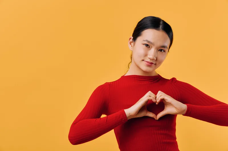 Young woman in red sweater making heart symbol with hands, smiling gently on orange background. Portrait of friendly female showing love gesture and positive emotions indoors.