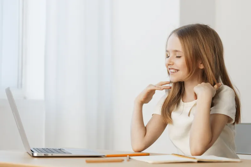 Joyful young girl with long hair enjoying online learning at home, dressed in a light top and smiling cheerfully, laptop and notebook in a bright environment