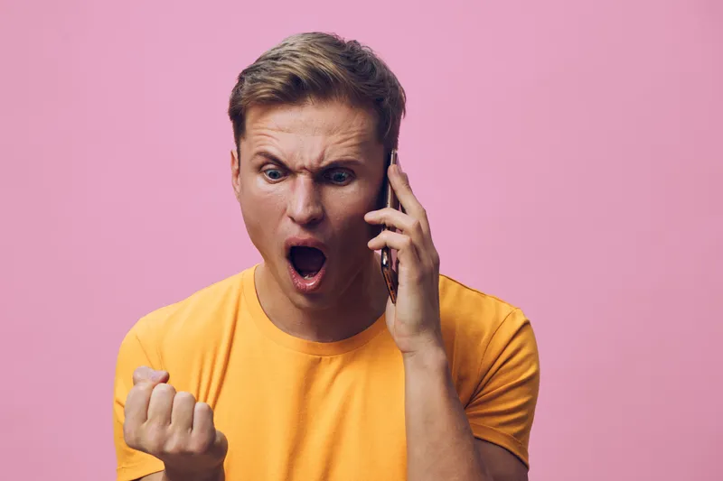 Close-up of a surprised young man with short brown hair, wearing a bright yellow t-shirt, shouting with wide eyes and clenched fist, talking on mobile phone on pink background, studio shot. People emo