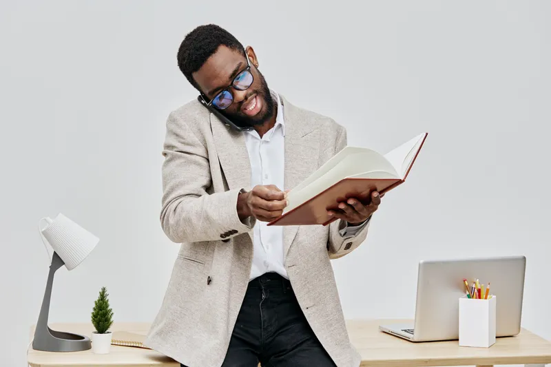 Joyful young African American man wearing glasses and beige blazer reading an open book at his workspace with a laptop, desk lamp, and plant, on a plain white background. People lifestyle concept.