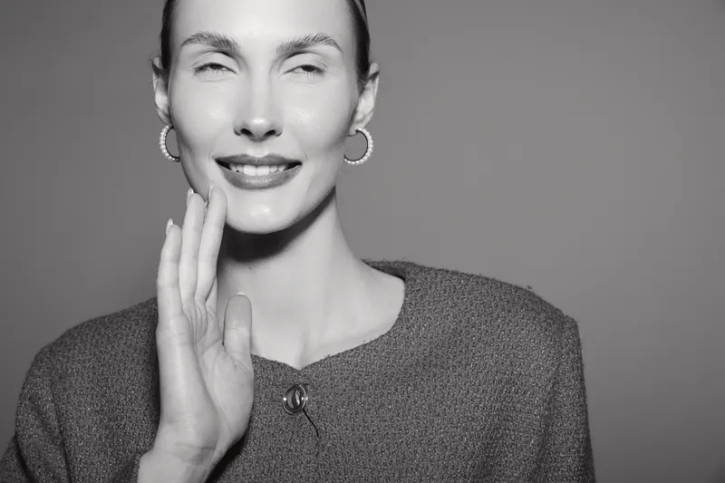 Smiling white woman with light skin and slicked hair touches her face in a studio portrait with soft lighting and gray background.