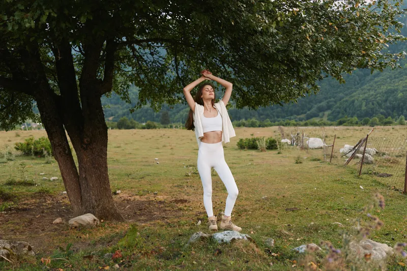 A young woman wearing white casualwear stands in a sunlit meadow beneath a large tree, striking a relaxed pose as calm countryside surrounds her.