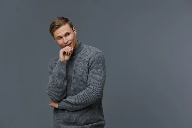 Smiling man, casual sweater, thoughtful mood, light brown hair, blue eyes, gray background, studio portrait, joyful expression, leisure, relaxed style, everyday fashion.