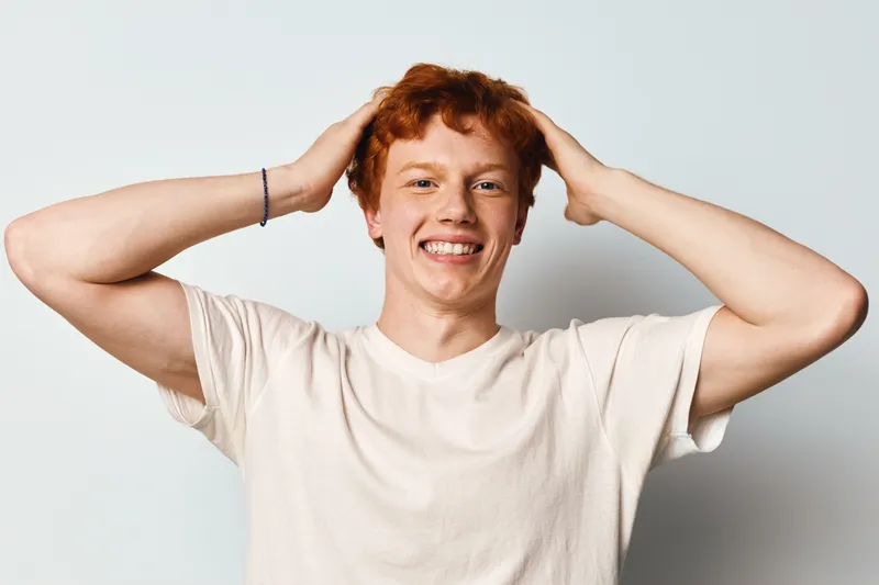 Young man with red hair smiling and holding head with hands wearing white t shirt against plain background. Casual confident person, cheerful, relaxed and happy expression portrait.