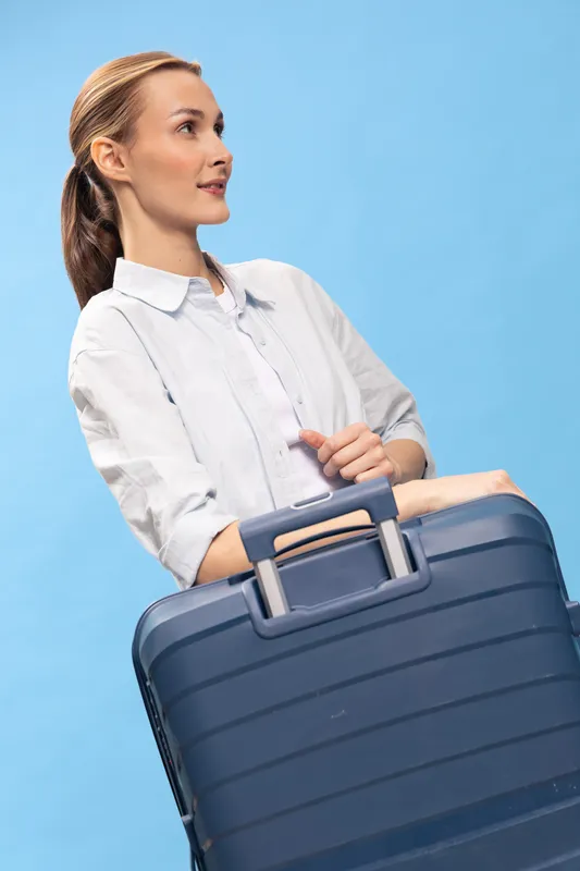 Young woman in white shirt holds dark blue suitcase, looking sideways on light blue background. Concept for travel, vacation, and business trip preparation.