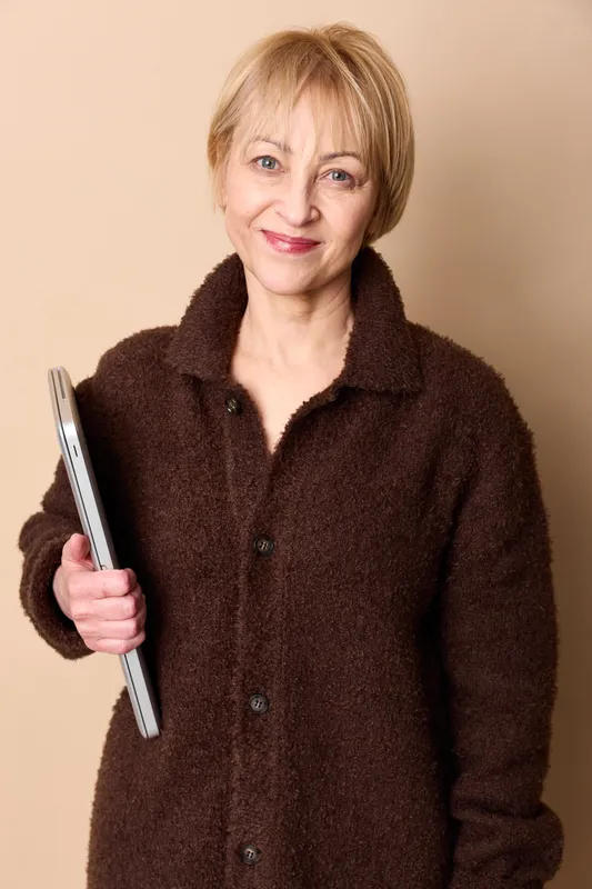 Elderly woman smiling in studio portrait, holding a tablet device at chest level. Beige background, soft light, calm confident mood. Concept for senior technology, communication.