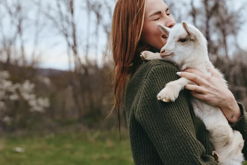 A woman in a green sweater cradles a young goat, her smile warm as they share a tender moment outdoors. Lifestyle scene emphasizing bond between human and animal in nature.