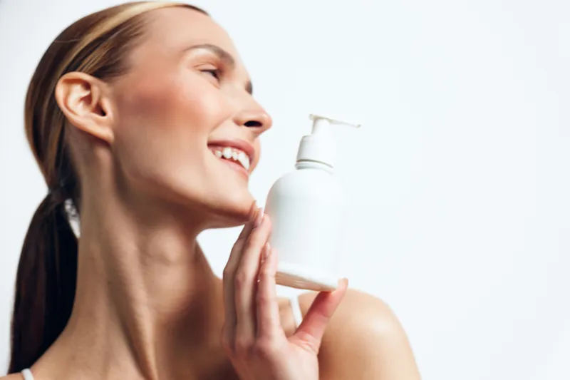 A smiling young woman with clear skin holds a white cosmetic bottle near her face against a plain white background. The image represents skincare, beauty, and personal care concepts.