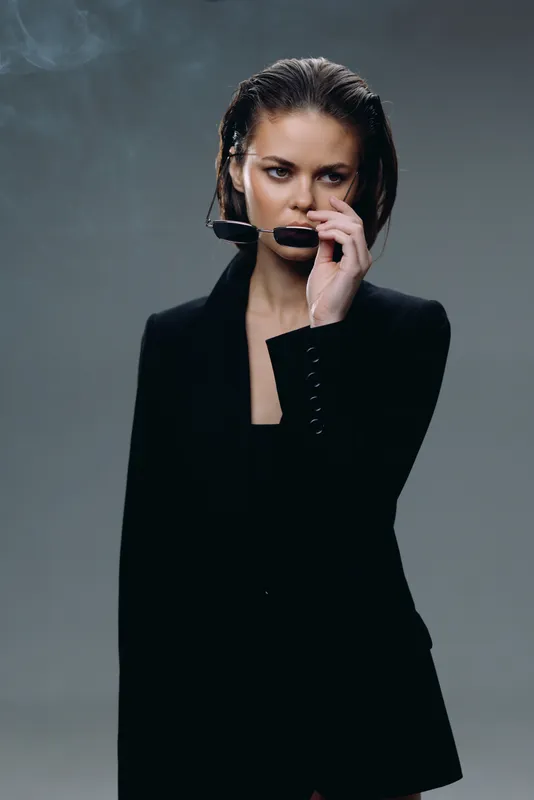Stylish woman in black blazer holding sunglasses with smoky background, confident and mysterious mood