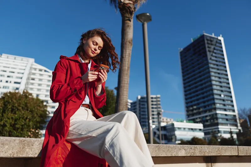 Young woman in a red coat sits on an urban ledge scrolling her phone, with palm trees and tall buildings behind, a candid city moment under bright light