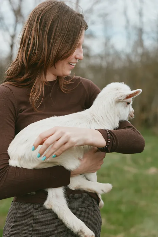 A woman outdoors holds a white goat kid, smiling softly in a rural field. The image captures gentle bonding, pastoral lifestyle, and affection for animals in a sunny countryside scene.