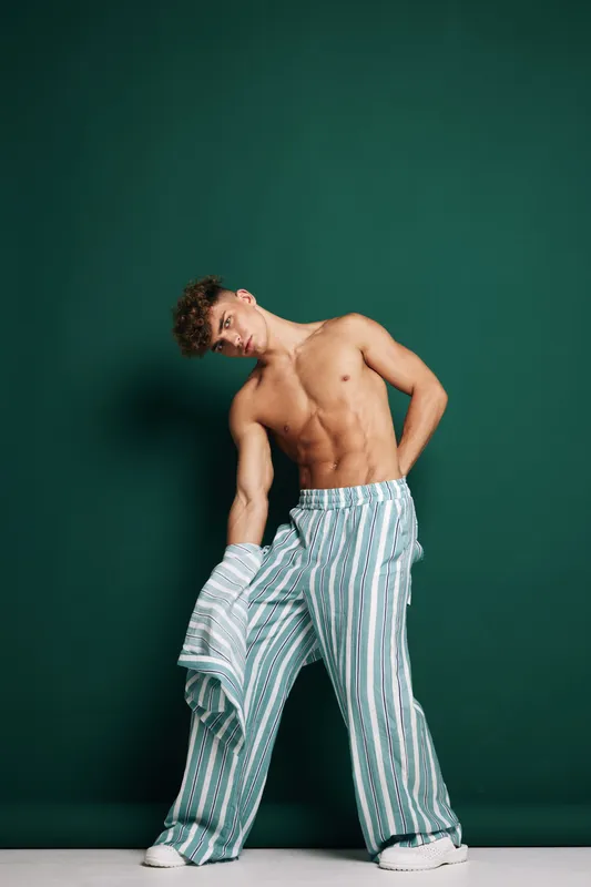 Young man with curly hair standing on white floor, wearing striped loose pants and holding matching shirt against teal background. Summer casual style with relaxed fashionable pose.