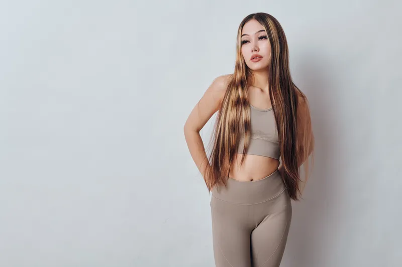 Young woman with long straight hair wearing beige knitted crop top and matching pants standing against plain light background in casual stylish outfit for fashion or lifestyle.