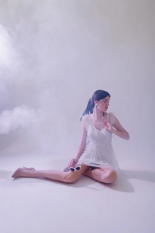 Young woman in a white lace dress sitting on the floor with sunglasses in hand. Soft lighting and mist create a dreamy fashion and beauty concept.