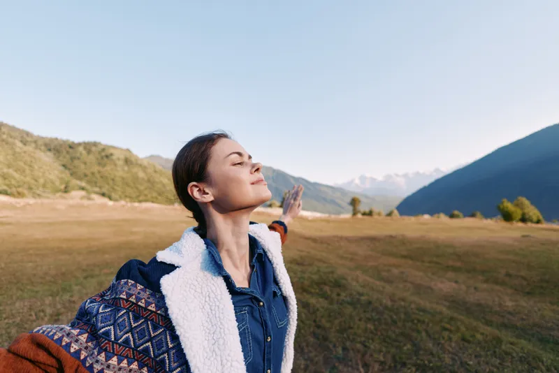 Woman in nature on mountains meadow with arms open, freedom and travel spirit outdoors portrait smiling and breathing fresh sunlight in peaceful countryside landscape.