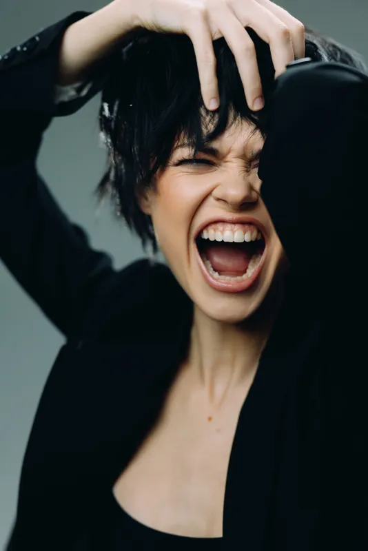 expressive woman shouting with hand in hair in dark outfit against blurred background