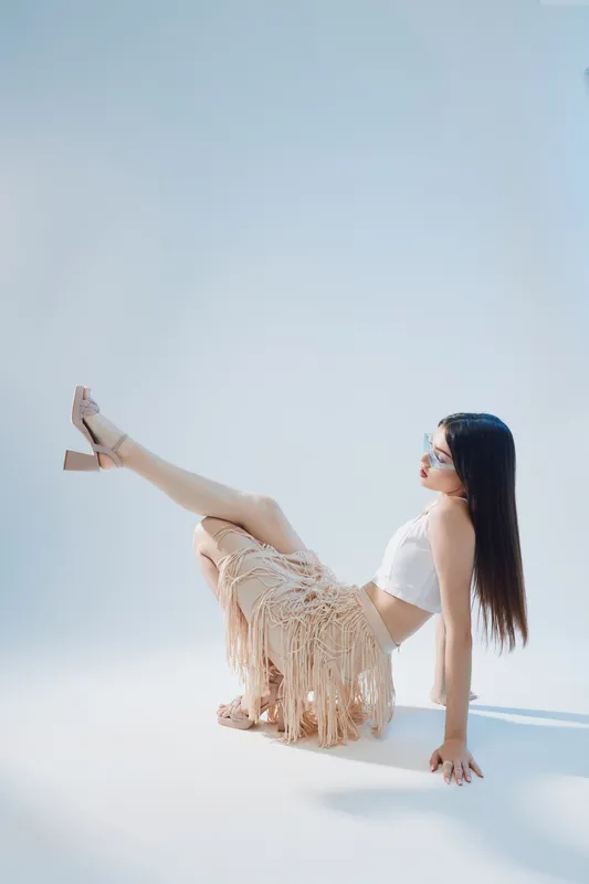 Young woman in white top and beige fringe skirt poses on floor with legs raised. Studio shot on light background. Concept of woman beauty fashion and elegance.