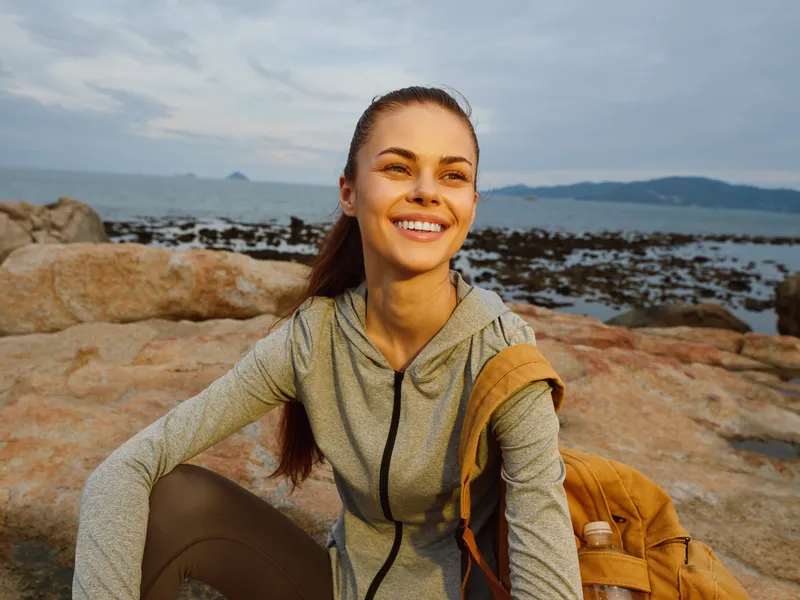 Smiling young woman in athletic wear on rocky beach at sunset, showcasing healthy lifestyle and outdoor fitness concept