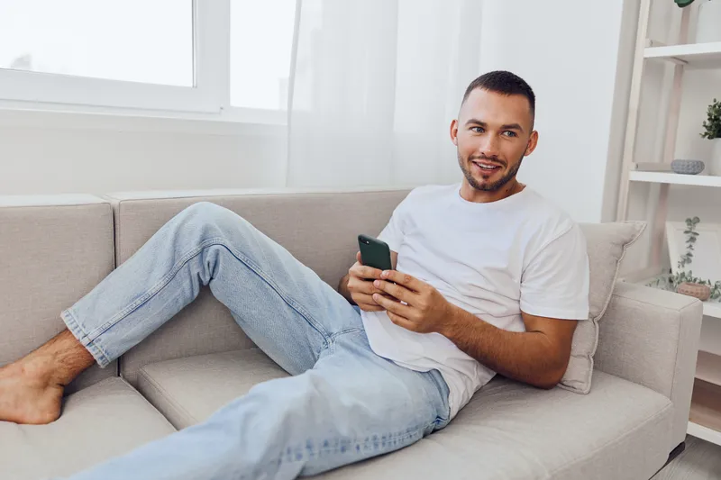 Relaxed man sitting on a couch, using a smartphone and smiling, showcasing modern lifestyle and digital connectivity in a cozy home environment.