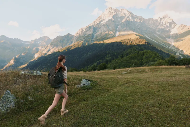 Young woman with a backpack hikes through a grassy mountain meadow under a bright sky, enjoying the peaceful natural landscape and fresh air.