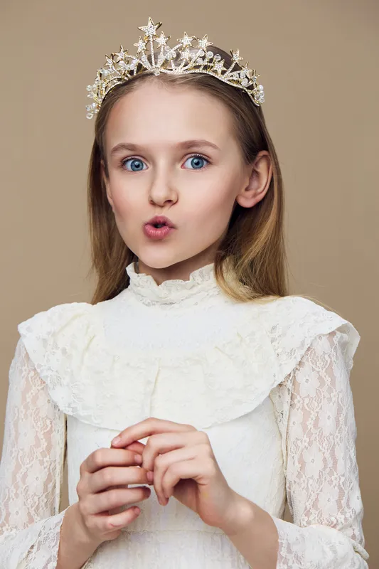 Young girl with a crown in a vintage lace dress, expressing surprise against a soft beige background, highlighting innocence and elegance in childhood fashion