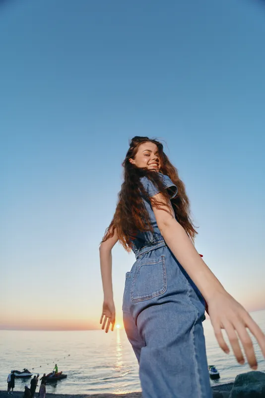 smiling woman in denim outfit enjoying sunset by the sea with long hair and natural light creating warm, joyful atmosphere near calm water horizon