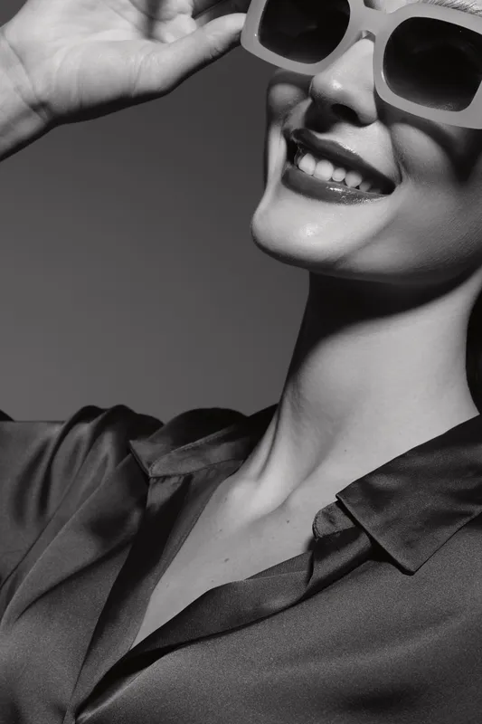Close-up of a smiling young Caucasian woman with fair skin wearing stylish sunglasses and a dark shirt in black and white photography.