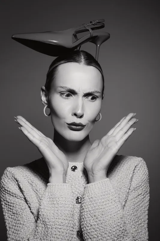 Young white woman with light skin wearing high heel on head looks confused with hands near face, studio portrait in black and white.