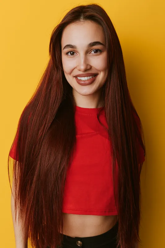 Young woman with long straight hair and red crop top smiling at camera on yellow background. Portrait of happy female with natural makeup and casual style in studio lighting.