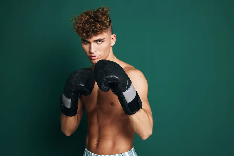 Boxer young man with curly hair positioned in fighting stance wearing black boxing gloves against green studio backdrop in athletic sports training session preparing for boxing match.