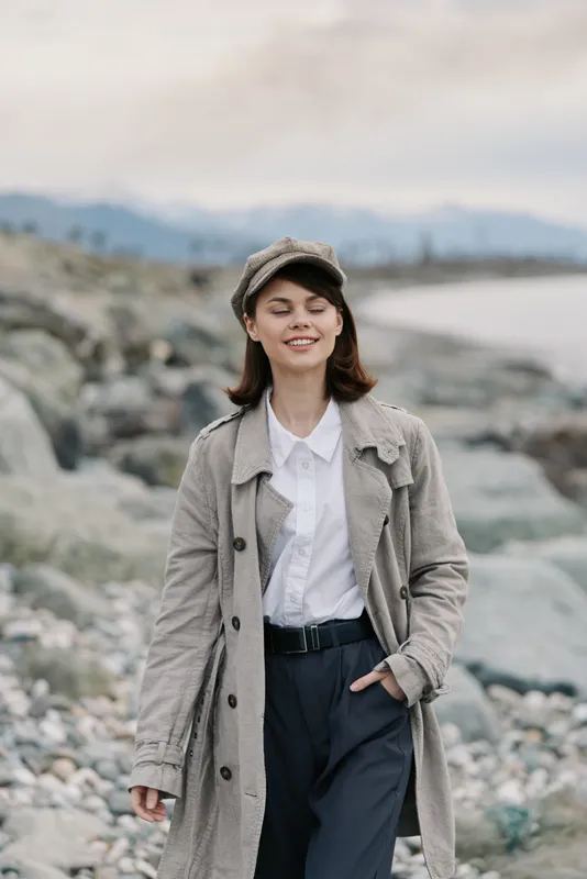 Young woman in a long coat and cap stands on a rocky beach with closed eyes, enjoying the calm atmosphere and natural landscape behind her. Concept of relaxation and nature.